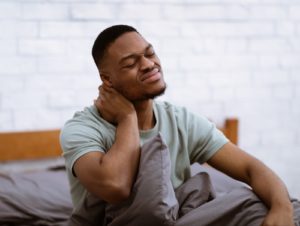 A man is sitting up in his bed, massaging his neck with his eyes closed in pain.