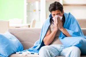 A man is sitting on the sofa wrapped in a blanket blowing his nose next to a pile of used tissues and a pillow.