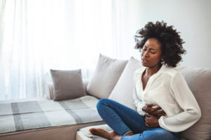 An African-American woman in jeans and a white button-up shirt sits on a sofa in pain, wincing and holding her abdomen with her hands.