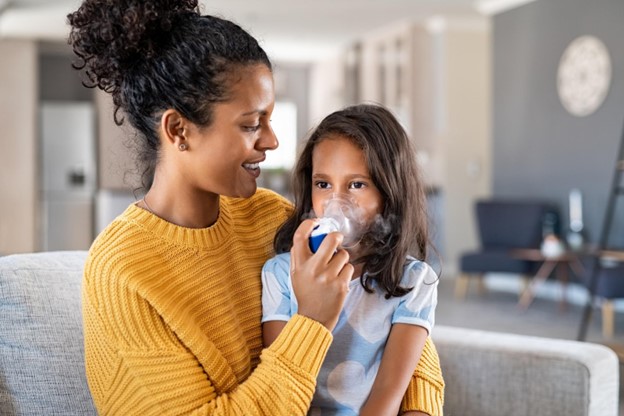 A mother in a yellow sweater is holding her daughter on her lap while administering an asthma breathing treatment using a nebulizer and face mask.