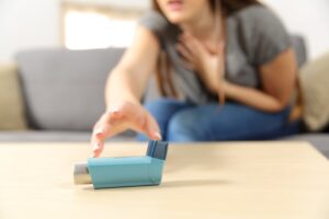 A focused shot of a woman sitting on the sofa, gripping her chest as she reaches for her asthma inhaler.