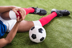 A close-up of an injured male soccer player sitting on the field, holding his knee with his hands near a soccer ball.