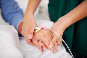 The hand and forearm of a woman lying in a bed are visible. She has an IV in place, and a nurse is holding her hand.