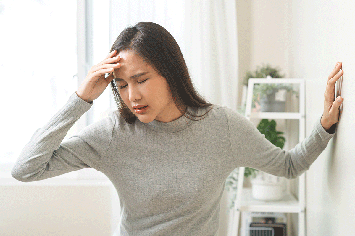 A woman with brown hair and a gray sweater holds one hand to her forehead and steadies herself against a wall with the other. Her eyes are closed.