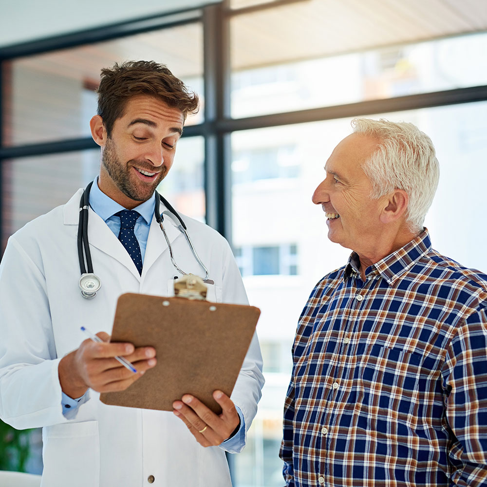 A man wearing a lab coat and holding a clipboard smiles and talks to an older man in a plaid shirt.