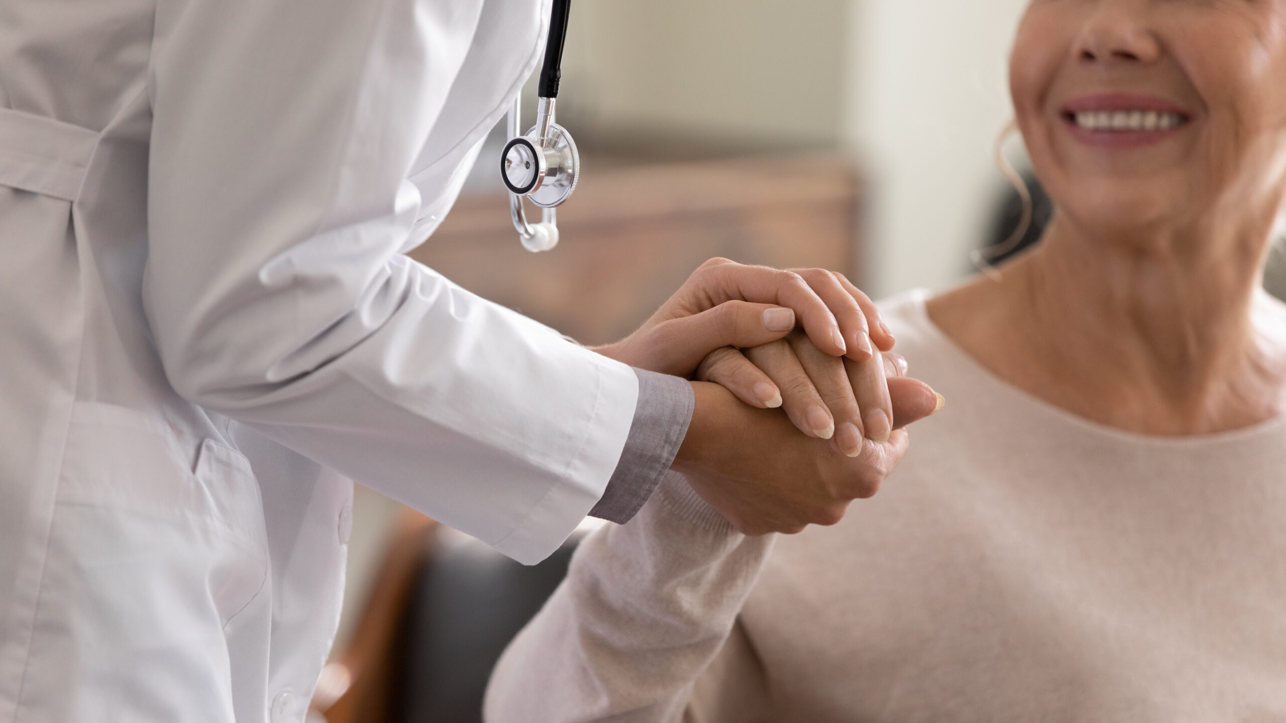 A woman in a white lab coat and a stethoscope holds the hand of an older woman wearing a sweater, who smiles up at her.