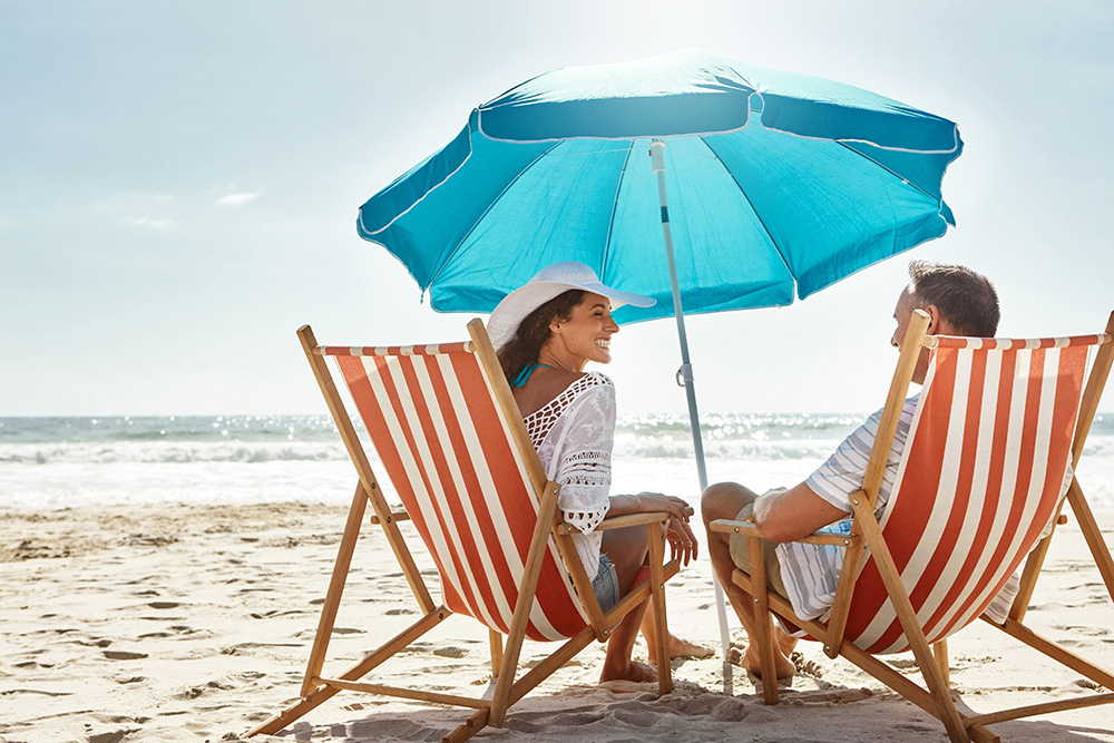 A man and a woman sit on striped beach chairs under a blue umbrella on a sandy beach.