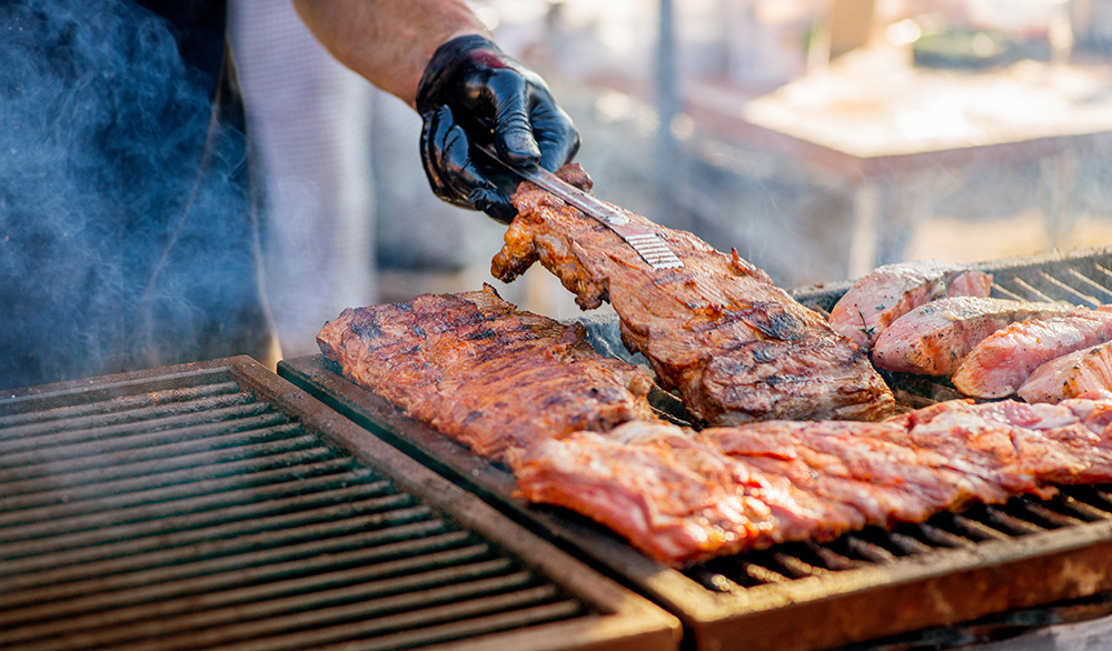 Person wearing gloves flips barbecue ribs on a charcoal grill during an outdoor backyard cookout.