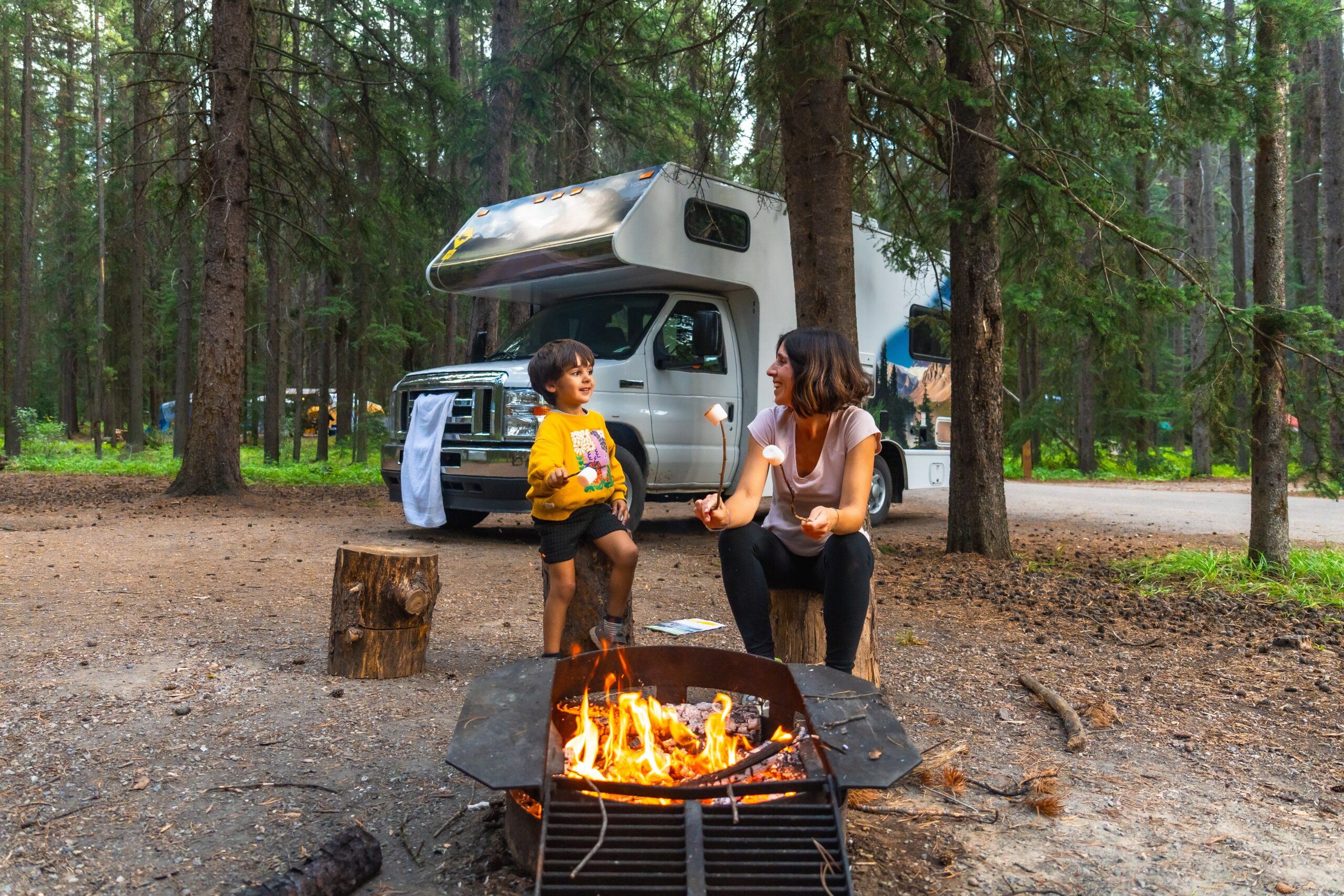 Mom and young son roasting marshmallows on sticks around campfire beside white camper van within a forest